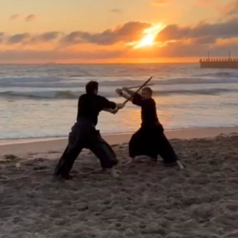 Two people practising Katori Shinto Ryu on a beach at sunrise.